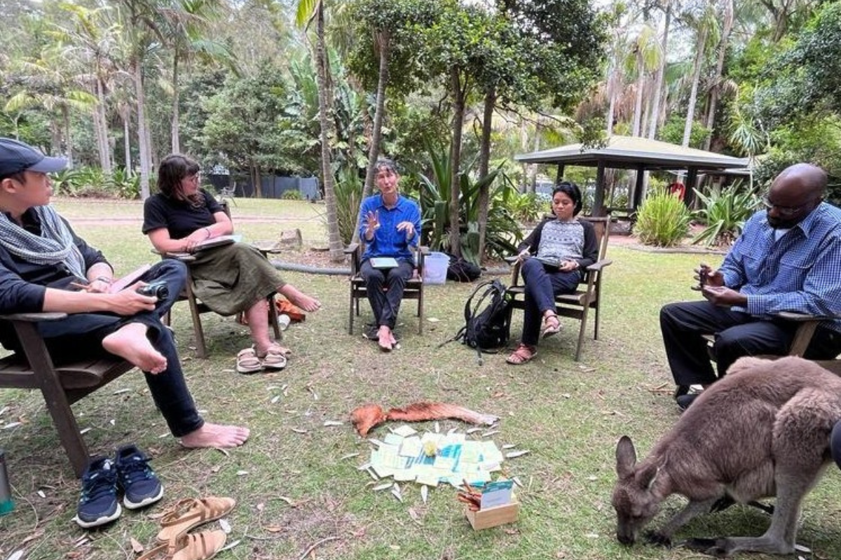 Group of six people seated in a circle outdoors, engaged in discussion, with a kangaroo grazing nearby and cards on the ground.